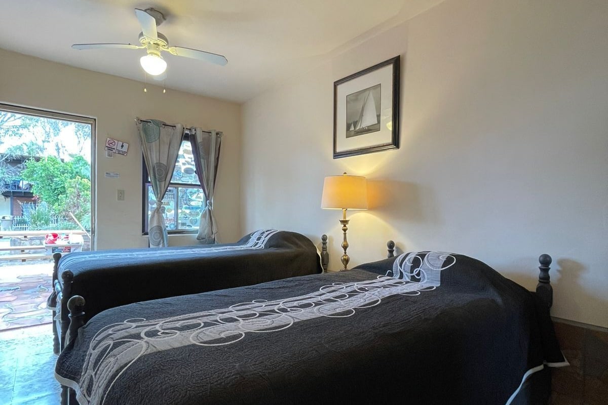 Une chambre d’hôtel avec deux lits, un ventilateur de plafond, une fenêtre avec des rideaux, un tableau encadré au mur et une lampe.