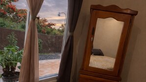A bedroom with a bed, a wooden dresser, a window with curtains, and a view of a garden.