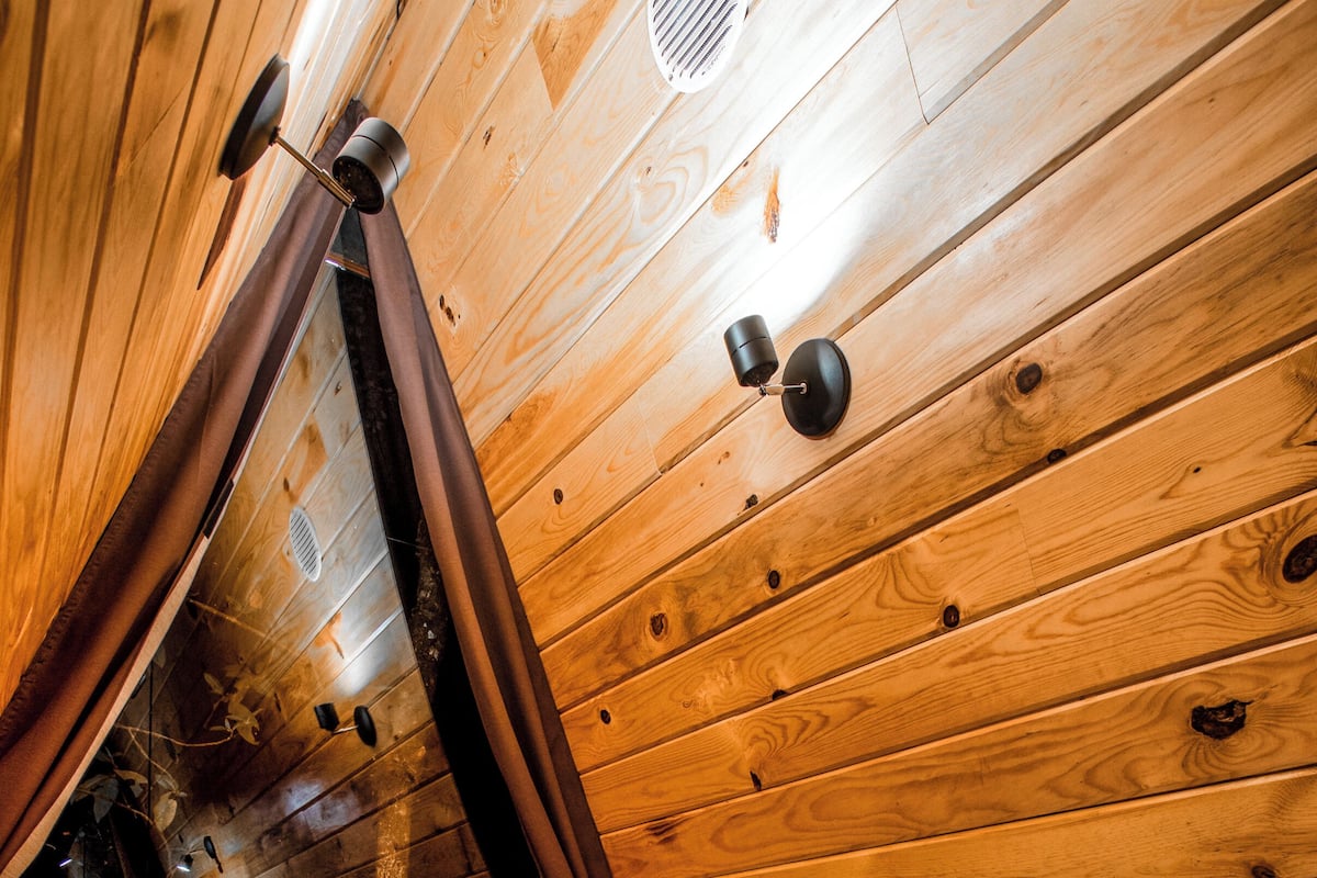 L’intérieur d’une cabane en bois, avec un lit, une fenêtre et une bouche d’aération au plafond.