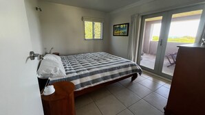 Um quarto com cama, mesa de cabeceira, cômoda, janela e porta que dá para uma varanda.