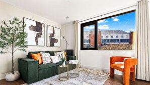 Ein modernes Wohnzimmer mit einem grünen Sofa, einem orangefarbenen Sessel, abstrakter Kunst, einem Topfpflanzen und einem großen Fenster mit Blick auf Gebäude.