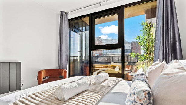 Ein modernes Hotelzimmer mit einem großen Bett, Blick auf die Stadt durch raumhohe Fenster und einem Balkon mit einer Pflanze.