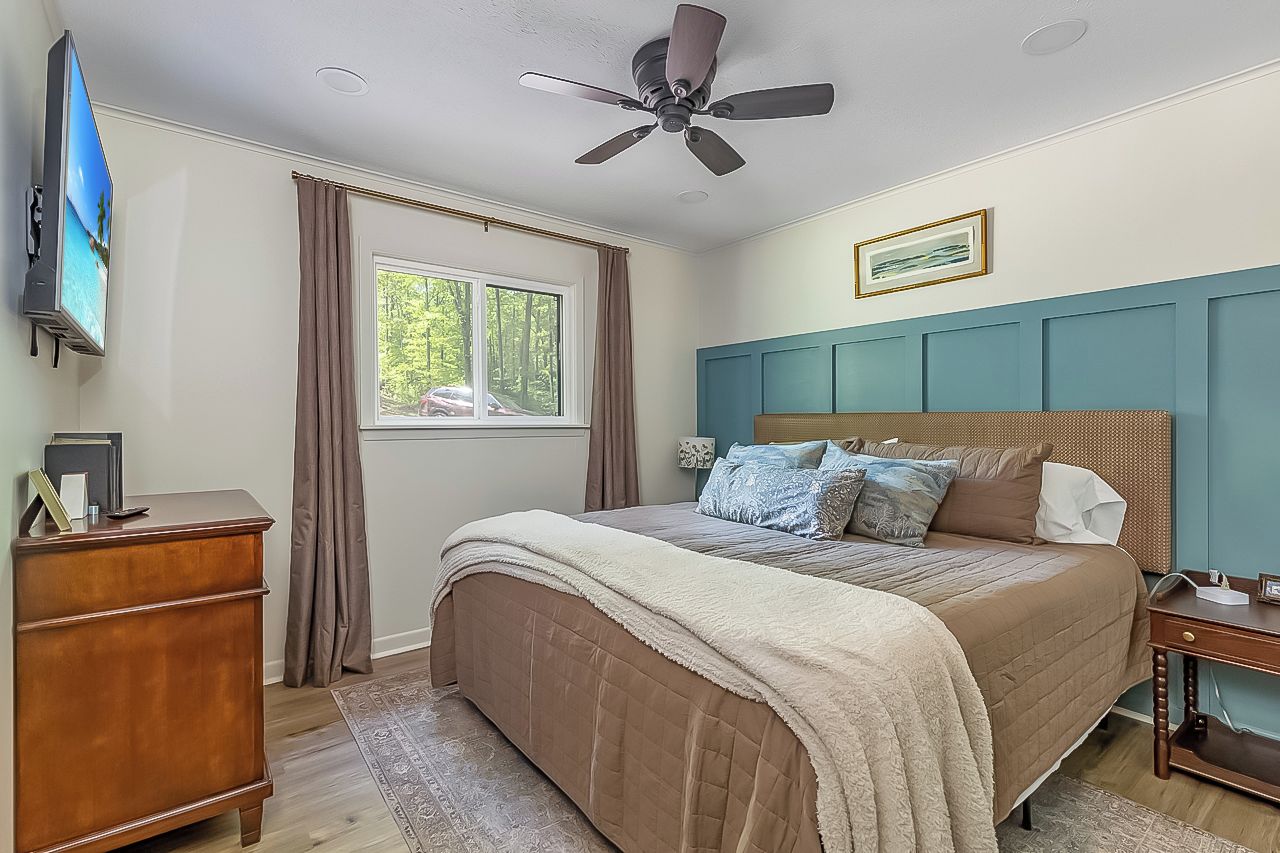 A bedroom with a bed, a ceiling fan, a dresser, a window with curtains, and a wall-mounted TV.
