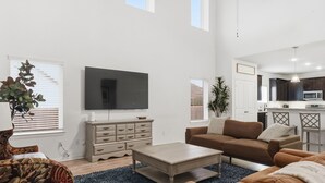 Ein modernes Wohnzimmer mit Flachbildfernseher, braunem Sofa, Couchtisch und Essbereich. Im Hintergrund ist eine Küche zu sehen.