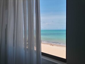 Een raam met dunne gordijnen, uitzicht op een strand en een heldere lucht.
