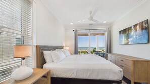 A hotel room with a bed, a desk, a lamp, a ceiling fan, and a large window with a view of the ocean.
