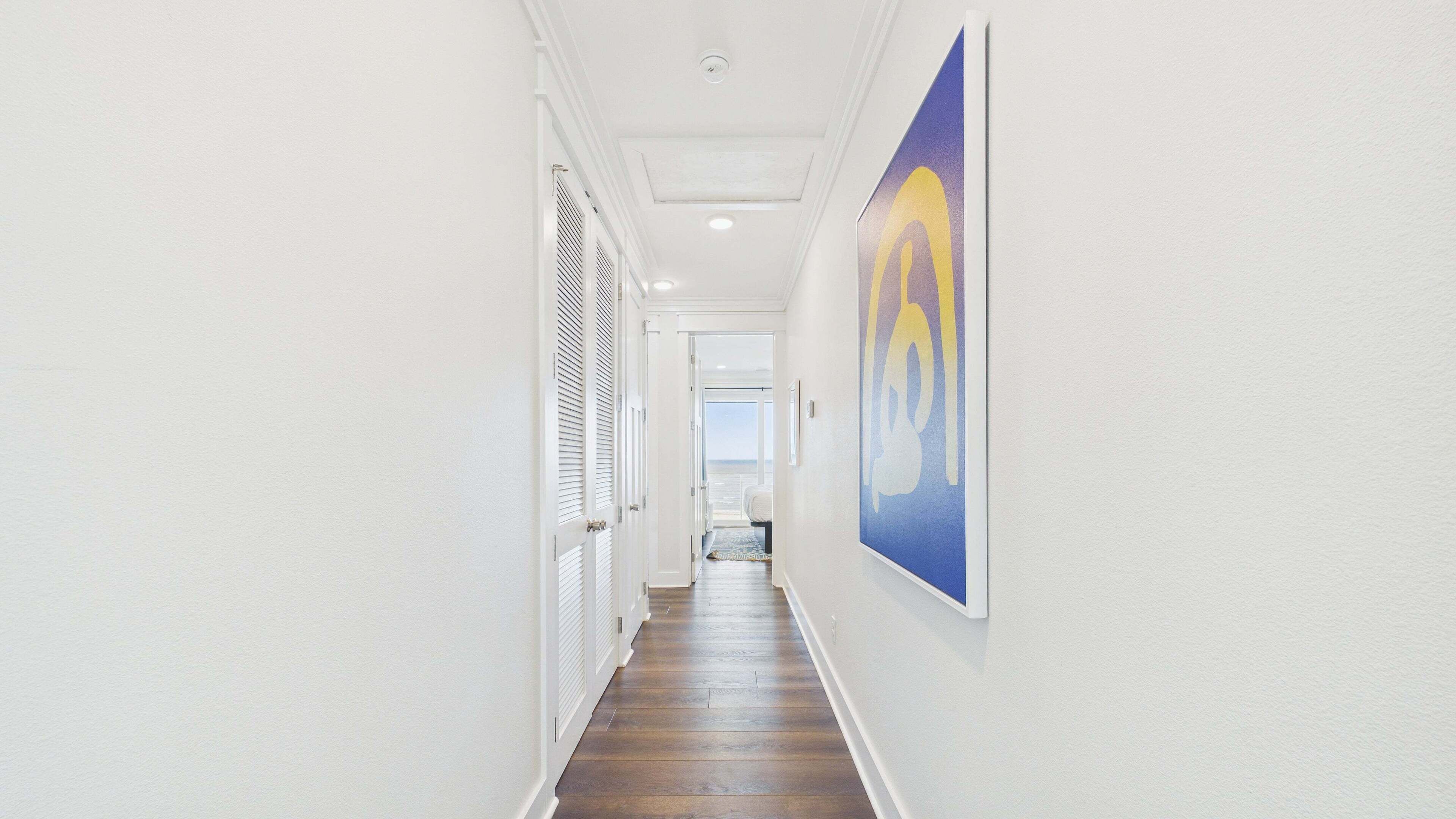A hallway with white walls, a wooden floor, and a large painting on the right wall.
