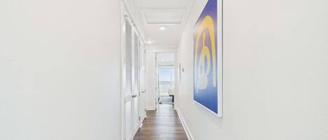 A hallway with white walls, a wooden floor, and a large painting on the right wall.