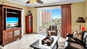 A living room with a television, a cabinet, a ceiling fan, a sofa, and a balcony with a view of buildings. - 🦩Book Me Baby! Pinkie's Island Oasis🦩 (Destin)