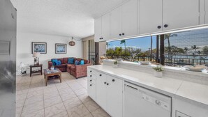 A modern kitchen with white cabinets, a large window, and a balcony with a view of palm trees. - New Listing! Steps to the beach in Kihei (Kihei)