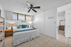 Ein Schlafzimmer mit Bett, Nachttischen, Deckenventilator, Fenster mit Jalousien, einer Tür und angrenzendem Badezimmer.