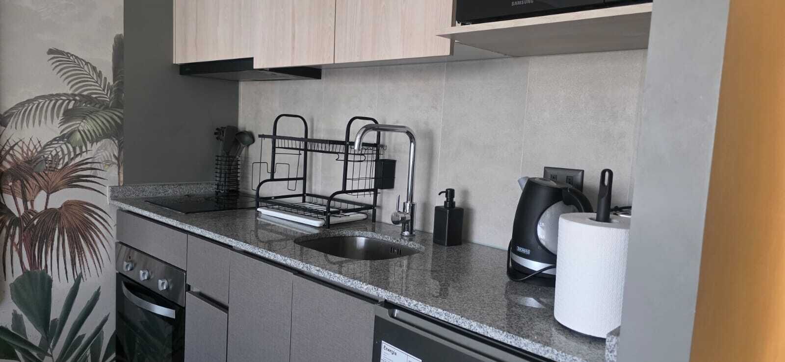 A modern kitchen with a stainless steel sink, black kettle, and a dish rack.
