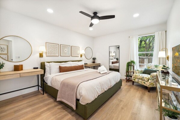 A bedroom with a bed, bedside tables, a desk, a chair, a ceiling fan, and a window with a view of trees.