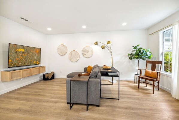 A modern living room with a grey sofa, a dining table, a TV, and a large window.