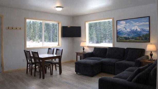A living room with a dining table, two couches, and a TV. There are windows with views of trees and mountains. - The Cabins at Riptide Resort (Seward)