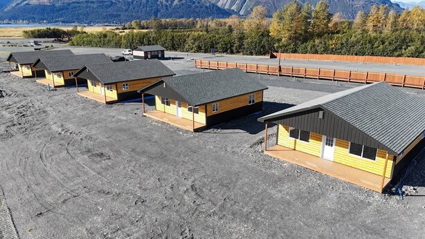 A row of small, wooden cabins with sloped roofs, arranged in a linear fashion on a gravel surface, with a forested mountain backdrop.