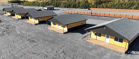 Une rangée de petites cabanes en bois, dotées de toits inclinés, disposées en ligne sur un sol de gravier, avec une montagne boisée en arrière-plan.