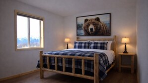 A bedroom with a wooden bed, two bedside lamps, a window with a view, and a bear painting on the wall. - The Cabins at Riptide Resort (Seward)