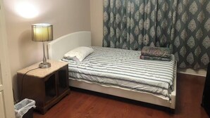 Un lit simple avec une couvre-lit à rayures, une lampe de chevet, une petite table de nuit en bois et un rideau à motifs.
