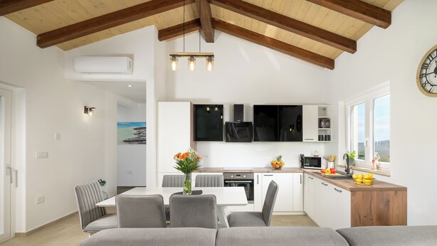 Un salon moderne avec un canapé gris, un coin repas, une cuisine avec des meubles blancs et un plafond en bois.