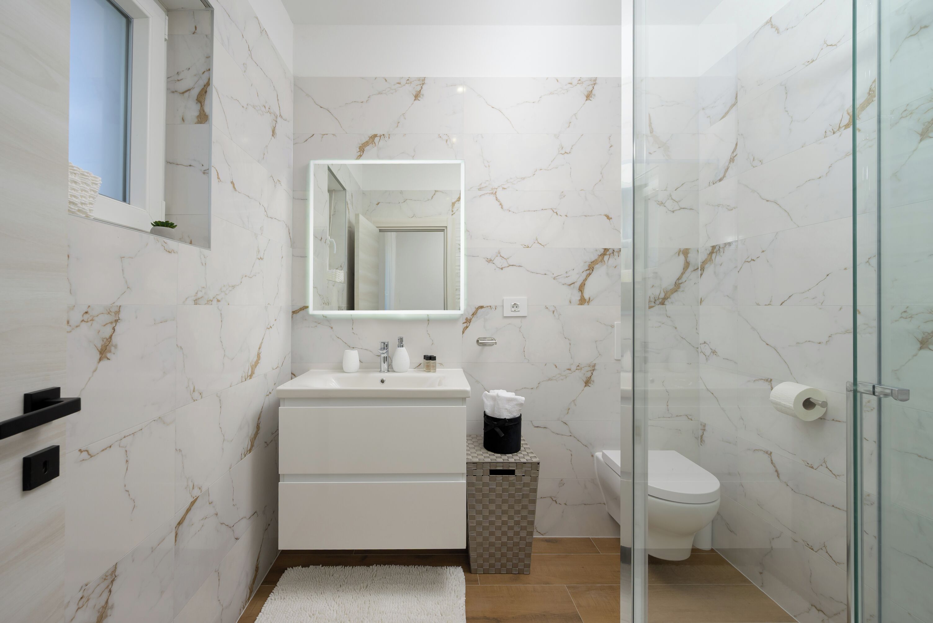 A modern bathroom with marble walls, a glass shower enclosure, a white sink, and a toilet.