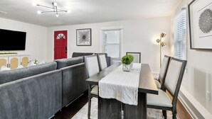 A modern living room with a gray sectional sofa, a dining table with chairs, a flat-screen TV, and a red door. - Spacious 3-Bedroom Uptown House – Steps to Streetcar & Downtown (New Orleans)