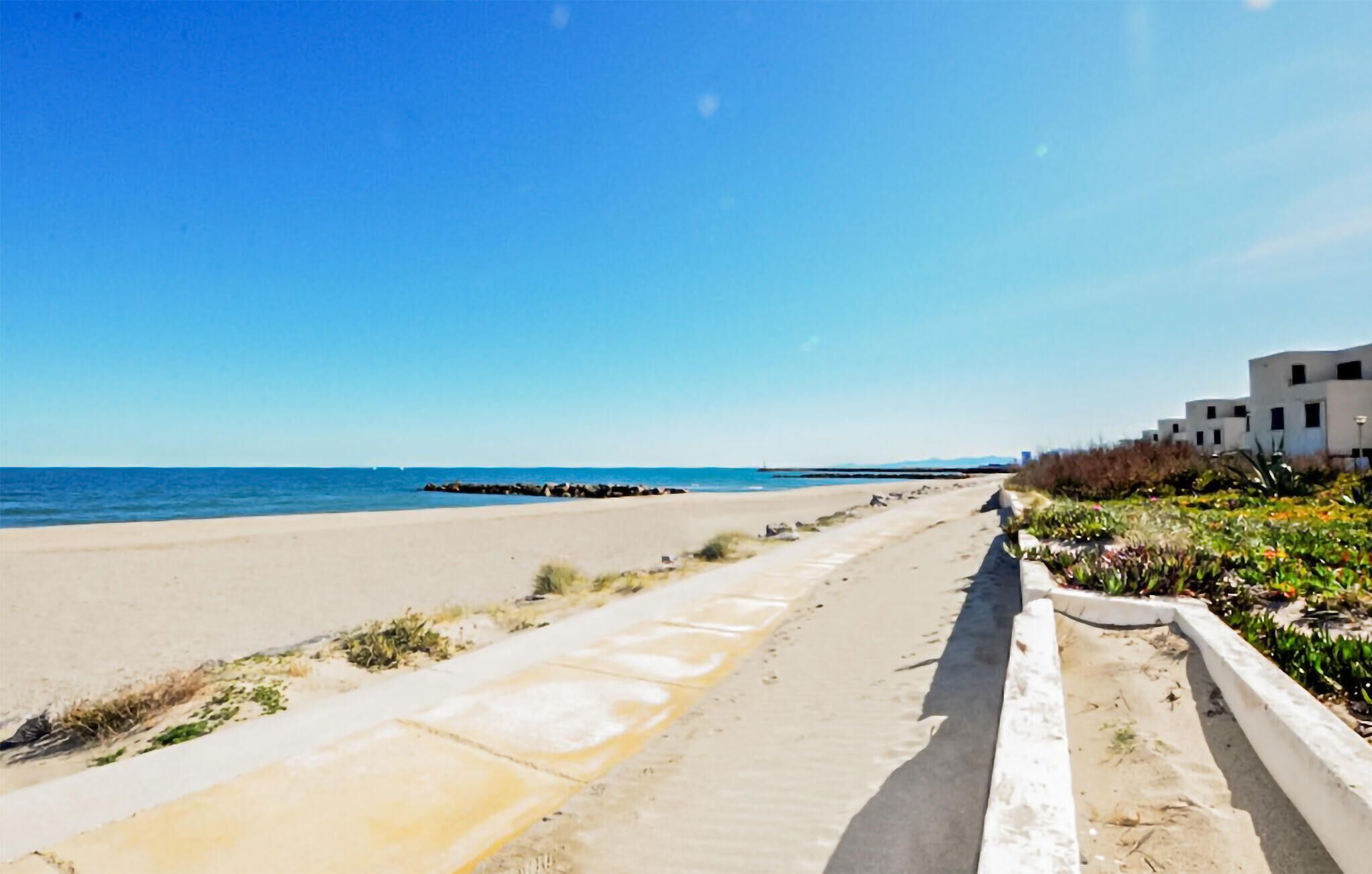 Une plage avec une promenade en béton, une digue rocheuse et des bâtiments modernes en arrière-plan.