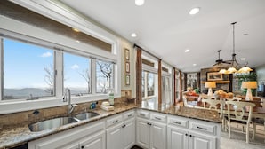 A kitchen with granite countertops, white cabinetry, a large window, and a dining area with a chandelier. - Panoramic Blue Ridge Views | Expansive Deck, Fireplace, One-Level Living (Blowing Rock)
