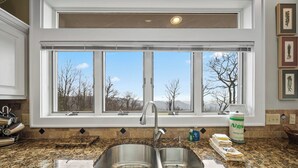 A kitchen with a double sink, a window with a view of trees and mountains, and a countertop with a paper towel dispenser. - Panoramic Blue Ridge Views | Expansive Deck, Fireplace, One-Level Living (Blowing Rock)