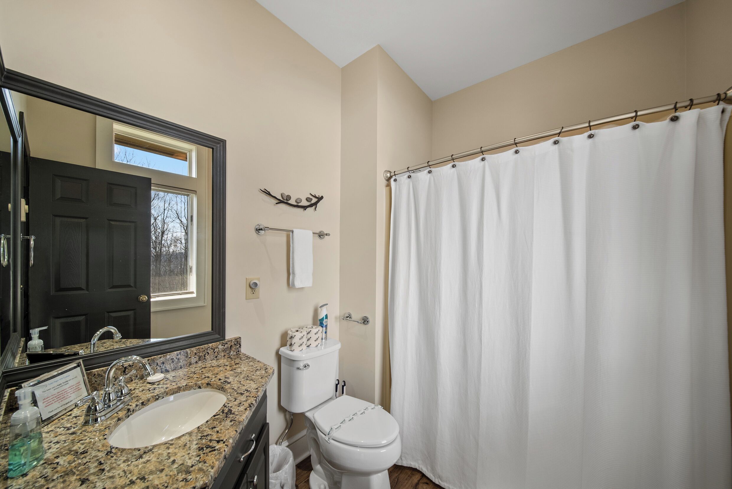 A bathroom with a granite countertop sink, a toilet, a shower curtain, a mirror, and a door leading to a window.