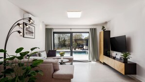 Ett modernt vardagsrum med en soffa, ett soffbord, en tv på ett träskåp och ett stort fönster med utsikt över en pool och utemöbler.