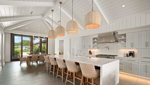 Une cuisine moderne dotée d’un grand îlot central, de luminaires suspendus et offrant une vue sur l’extérieur.