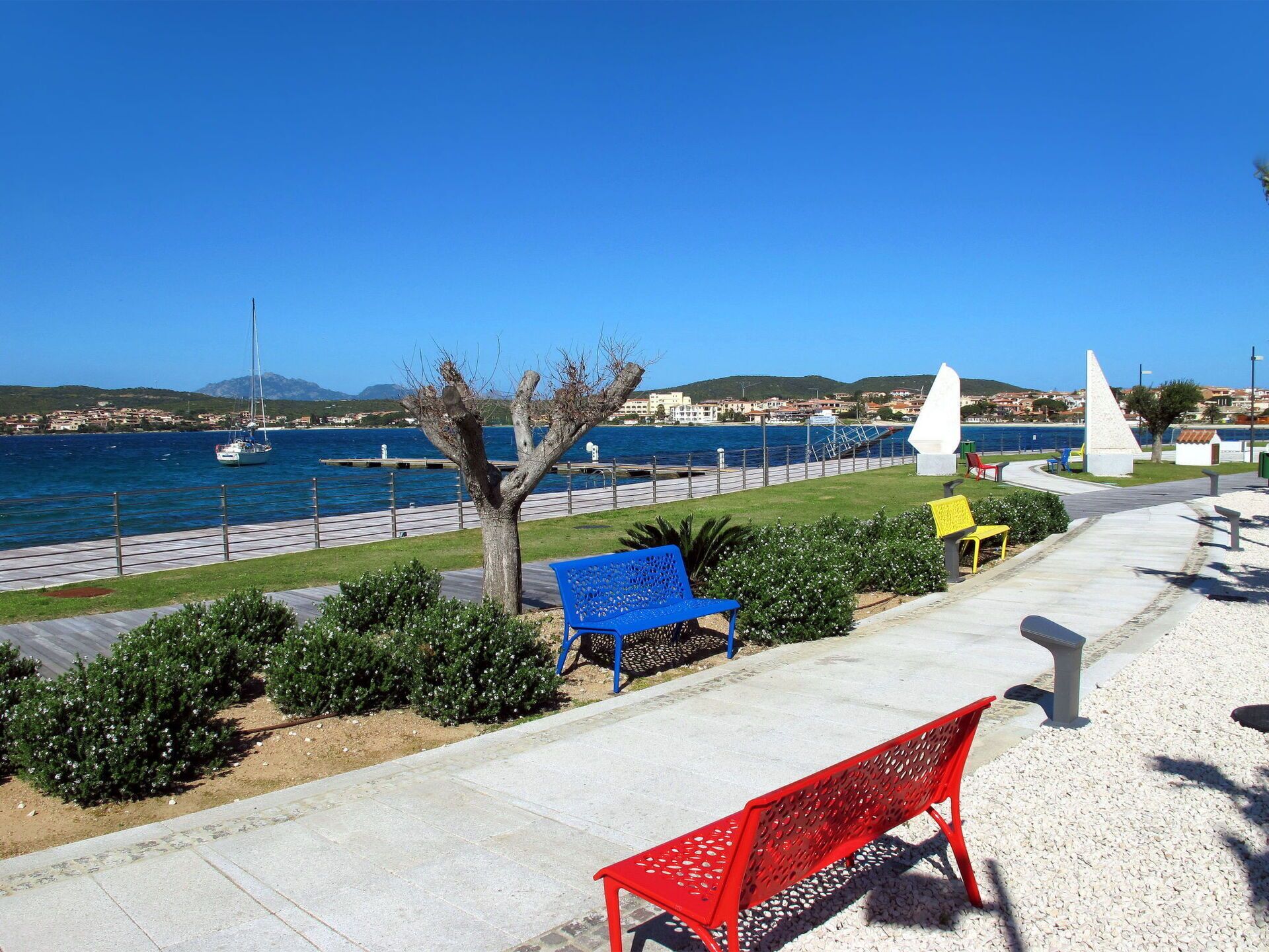 A coastal promenade with benches, a clear view of the sea, and a distant town.