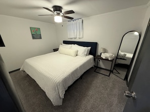 Un dormitorio con cama, ventilador de techo, mesita de noche con lámpara, espejo y un cuadro en la pared.