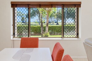 A room with a dining table, chairs, and a window with a view of a parking area and trees. - Esplanade Water-Front 2BR | Pool & Balcony (Cairns North)