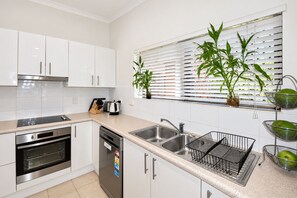 A modern kitchen with white cabinets, stainless steel appliances, a window with blinds, and potted plants. - Esplanade Water-Front 2BR | Pool & Balcony (Cairns North)