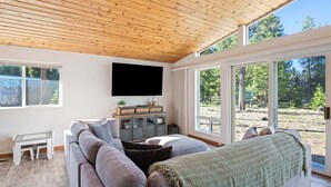En moderne stue med en stor krogsofa, et fladskærms-tv monteret på væggen og udsigt til et skovområde gennem store vinduer.