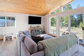 A modern living room with a large sectional sofa, a flat-screen TV mounted on the wall, and a view of a wooded area through large windows.