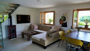 Ein modernes Wohnzimmer mit einer Couch, einem Couchtisch und gelben Sesseln. Ein Kamin, ein an der Wand montierter Fernseher und ein Fenster mit Blick auf Grün sind vorhanden.