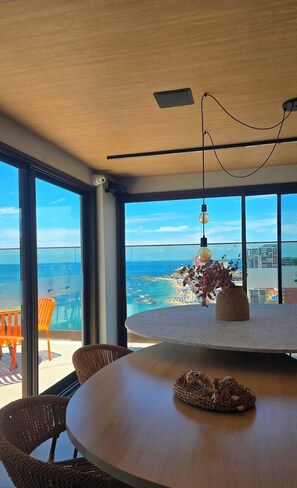 A modern dining area with a round table, wicker chairs, a hanging light fixture, and a view of the ocean and cityscape through large windows. - Studio with the best view of Salvador. (Salvador)