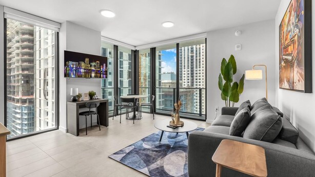 A modern living room with a sofa, coffee table, and dining area, large windows with city views, and a mounted TV. - Luxury High-Rise 1BR in Downtown Miami (Miami)
