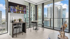 A modern living room with a desk, chairs, a coffee table, and a large window with a city view. - Luxury High-Rise 1BR in Downtown Miami (Miami)