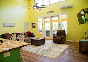 Un salon comprenant un canapé, des fauteuils, une table basse, une table à manger, un ventilateur de plafond, une grande fenêtre donnant sur un beau paysage et une porte menant à l’extérieur.