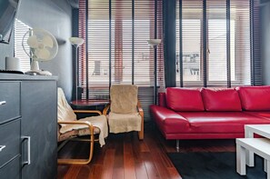 Ein Zimmer mit einem roten Sofa, einem Holzstuhl, einem kleinen runden Tisch, einem Schreibtisch und einem Schrank.