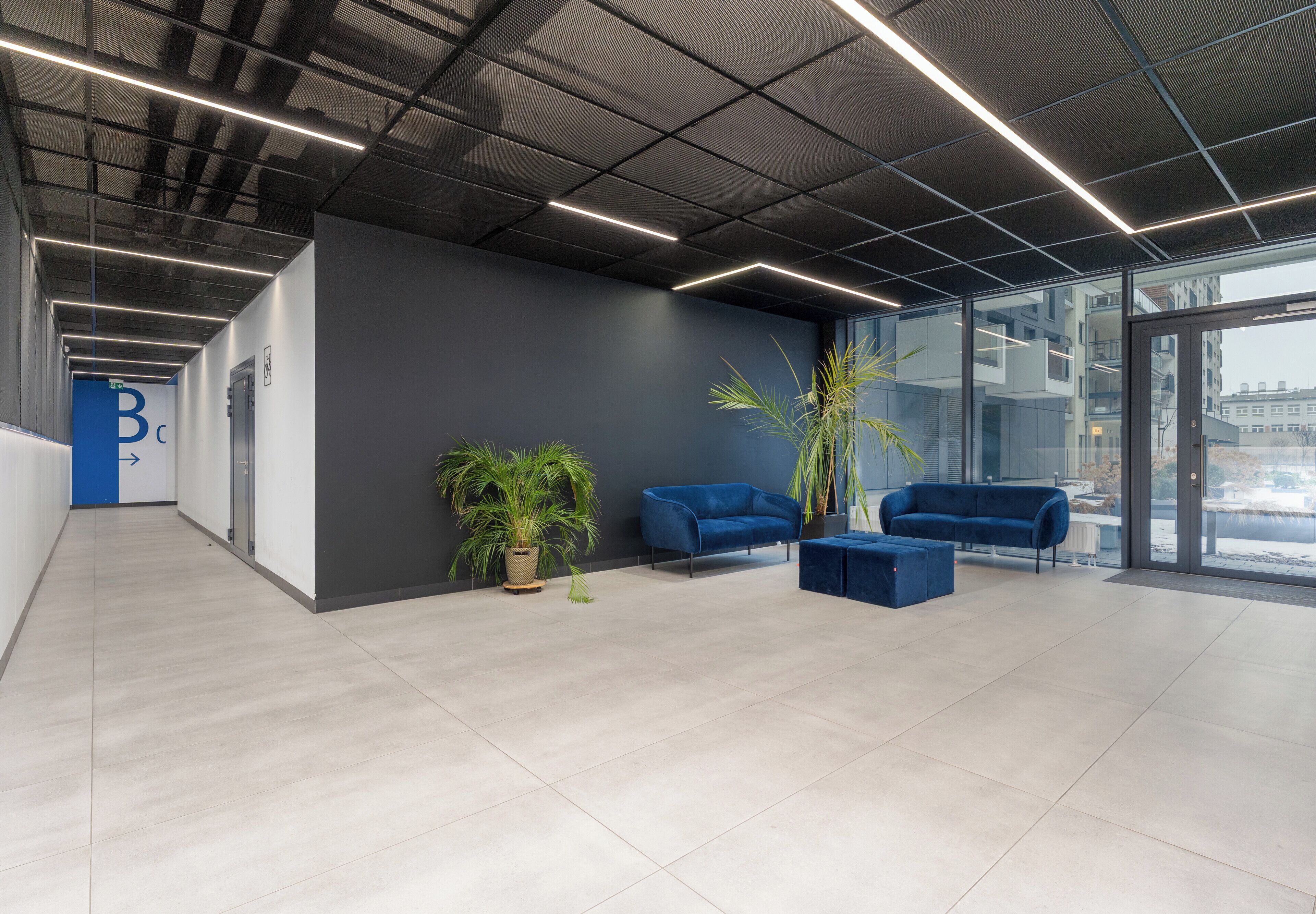 Un espacio de oficina moderno con un largo pasillo, sofás azules y plantas en macetas.