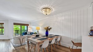 A dining area with a wooden table, white chairs, a chandelier, and a view of greenery outside. - New to VRBO, Oceanwalk, Near Beach, Outdoor and Indoor Pool, Hot Tub (Hilton Head Island)