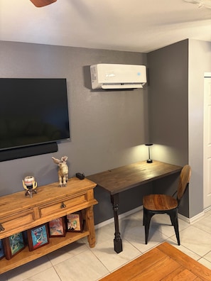 A room with a flat-screen TV mounted on the wall, a wooden desk with a chair, a small table with decorative items, and an air conditioning unit. - Studio Chalet Guesthouse in Show Low (Show Low)