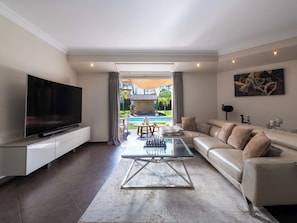 Ein modernes Wohnzimmer mit Flachbildfernseher, Glastisch, beiger Couch und Blick auf einen Swimmingpool und Außenbereich.
