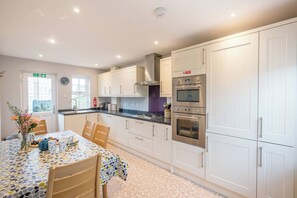A modern kitchen with white cabinets, a dining table with a colorful tablecloth, and a visible clock on the wall. - North Warren Cottage, Aldeburgh (Aldeburgh)