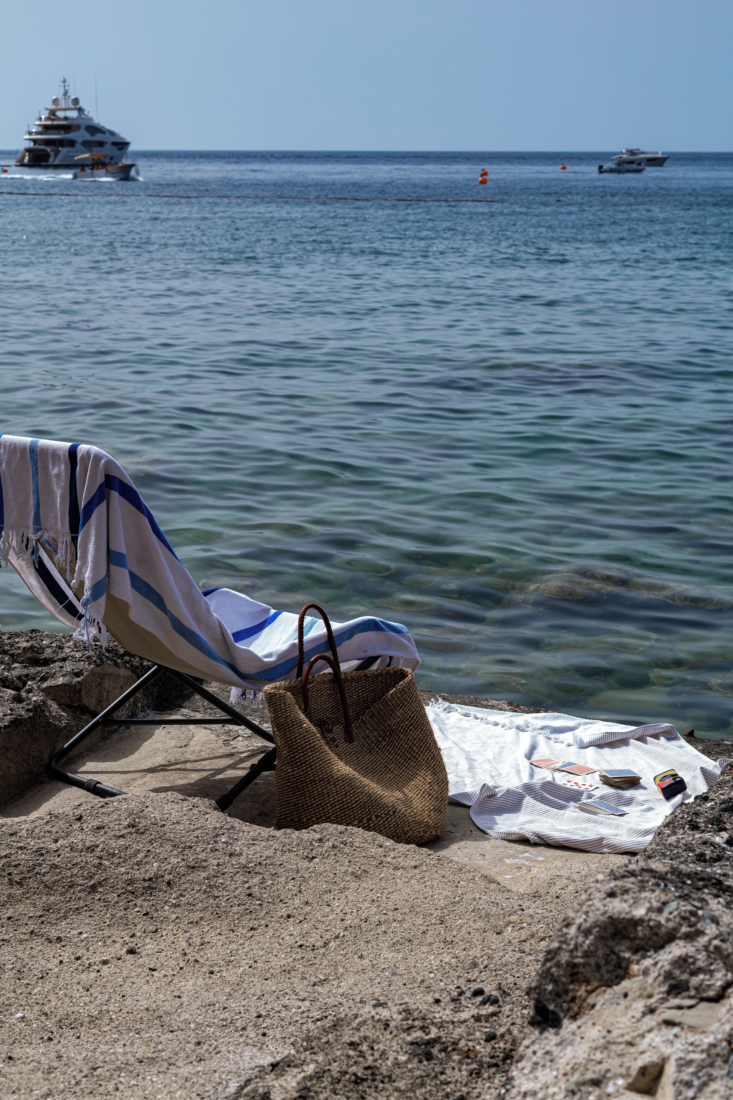 Una sedia da spiaggia con un asciugamano a righe e una borsa di vimini su una riva rocciosa, con vista su un mare calmo e barche in lontananza.